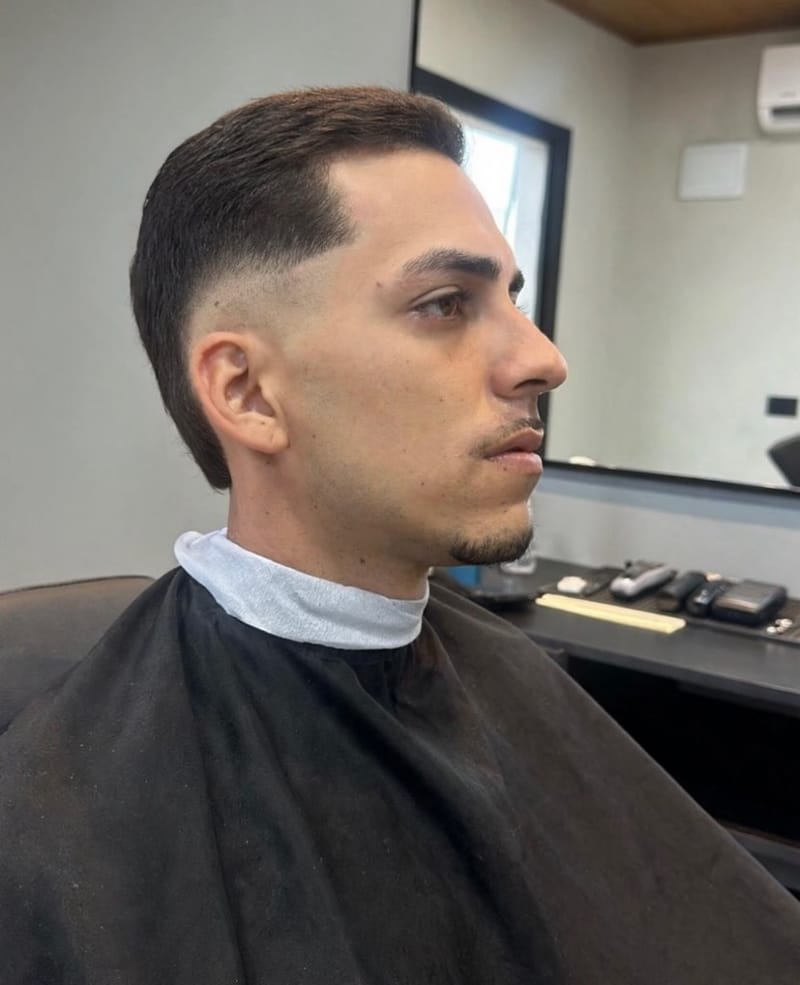 Cliente de perfil com cabelo curto penteado para trás e laterais raspadas em transição suave, sentado na cadeira da barbearia.