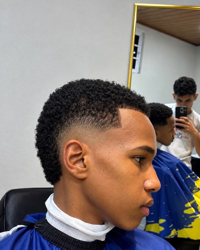 Jovem de perfil com cachos curtos no topo, laterais baixas e contorno marcado na testa.
